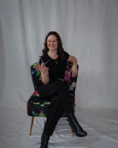 Meet Meredith Dellaria Salons Team | Dellaria Salons Meredith hair colorist and stylist with 15+ years experience on the Dellaria Salons team in Massachusetts holding professional shears in a floral accent chair